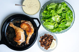 Two chicken thighs on a cast iron skillet with three bowls containing salad, caesar dressing, and pan fried croutons