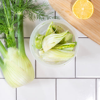 Sliced Fennel in a reusable container on a tile background, next to a large unsliced bundle of fennel